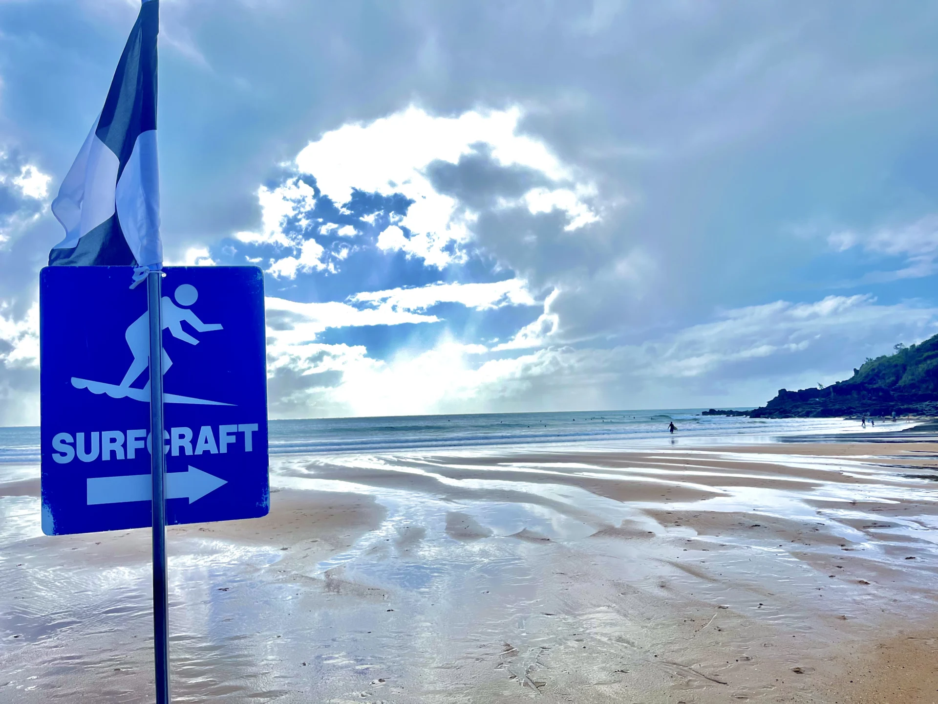 Beach sign pointing to surfcraft area at Agnes Water, with flag and distant surfers on a sunny day.